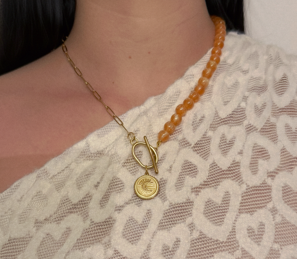 Collier asymétrique avec une moitié en chaîne dorée, une moitié en perles orange ambré, un fermoir T-bar à l'avant et un pendentif médaille lune et soleil.