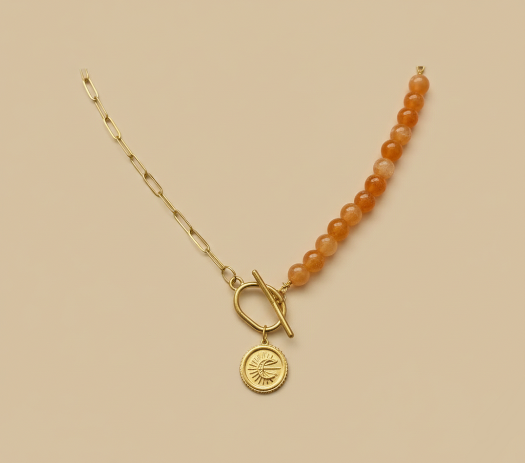 Collier asymétrique : moitié chaîne dorée, moitié perles orange, avec fermoir T-bar et médaille lune à l'avant, sur fond beige neutre.
