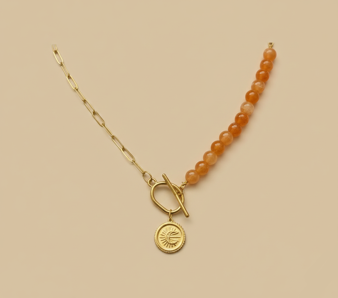 Collier asymétrique : moitié chaîne dorée, moitié perles orange, avec fermoir T-bar et médaille lune à l'avant, sur fond beige neutre.
