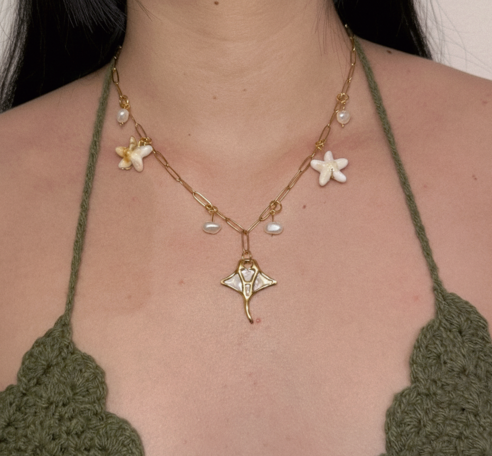 Collier doré d'inspiration marine avec un pendentif raie manta nacré, des étoiles de mer et des perles d'eau douce, porté sur un décolleté.