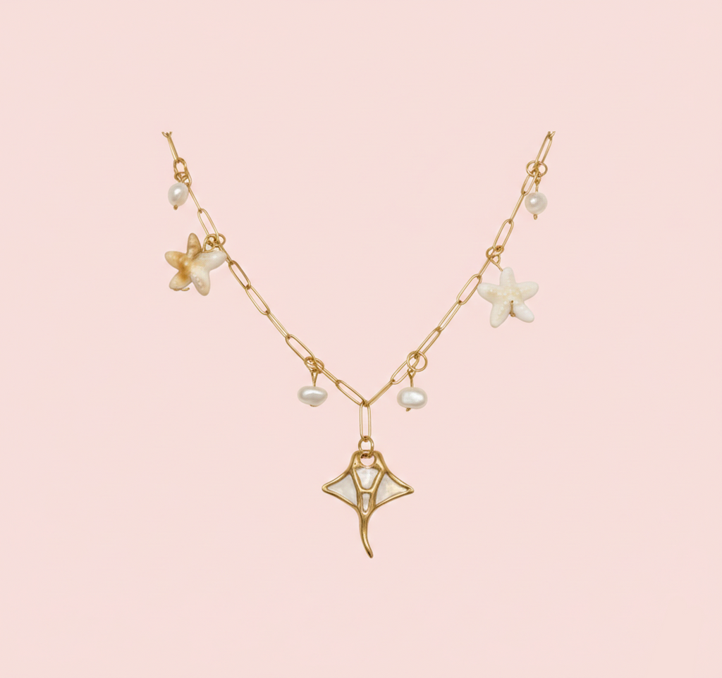 Collier doré à maillons rectangulaires avec un grand pendentif raie manta, orné d'étoiles de mer nacrées et de perles d'eau douce, sur fond rose clair.