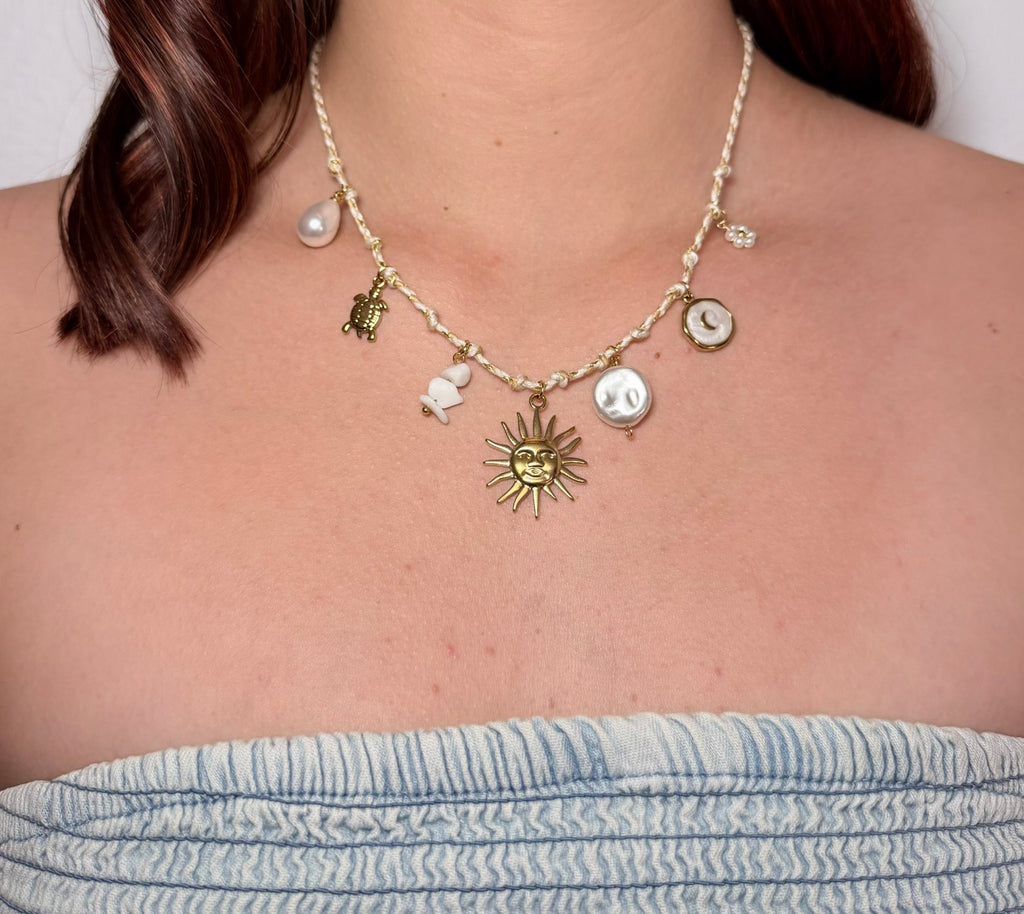 Gros plan sur un collier artisanal tressé blanc et doré, orné de breloques dorées et marines (soleil, lune, tortue, étoile de mer et coquillages), porté par une personne en bustier en jean.