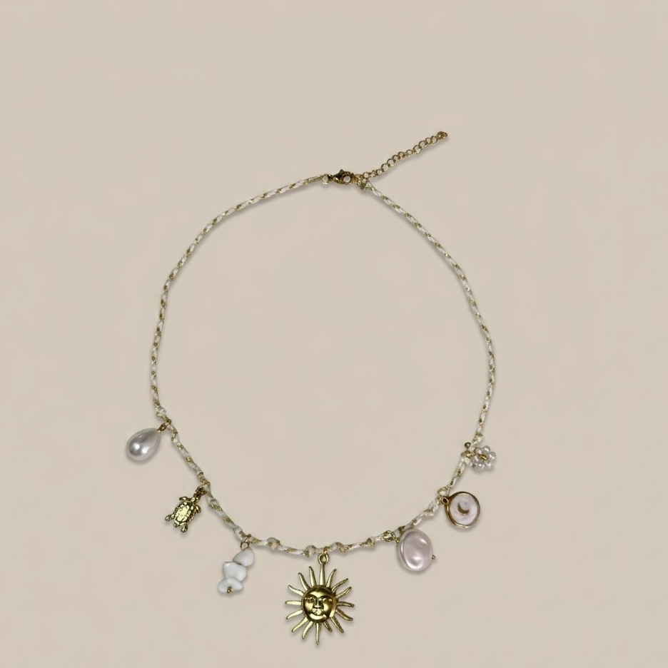 Gros plan sur un collier artisanal tressé blanc et doré, orné de breloques dorées et marines (soleil, lune, tortue, étoile de mer et coquillages) sur un fond beige clair.