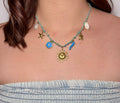 Gros plan sur un collier artisanal tressé bleu et vert, orné de breloques dorées et marines (soleil, lune, tortue, étoile de mer et coquillages), porté par une personne en bustier en jean.