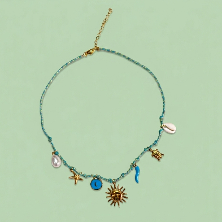 Collier artisanal tressé bleu et vert, orné de breloques dorées et marines (soleil, lune, tortue, étoile de mer et coquillages) sur un fond vert pastel.
