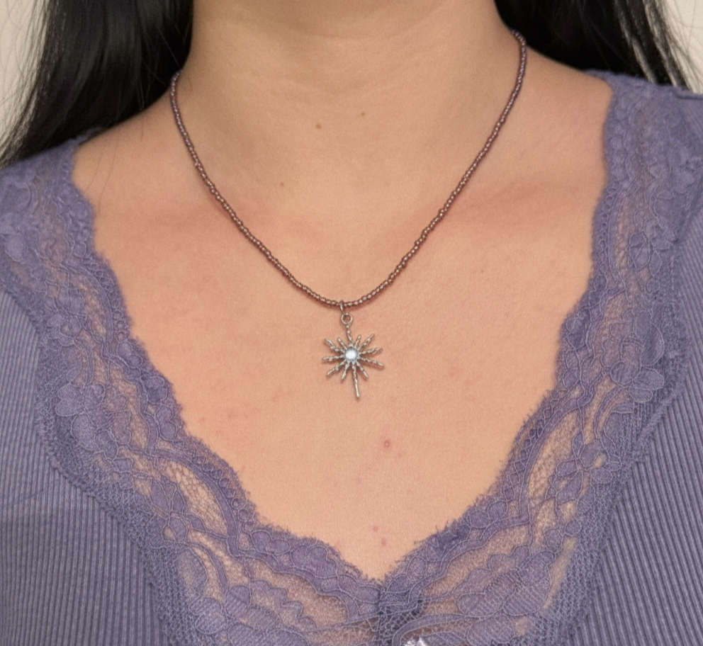 Collier en fines perles de couleur acrylique violette avec un pendentif en étoile d'argent stylisée, porté sur un décolleté avec un haut en dentelle.