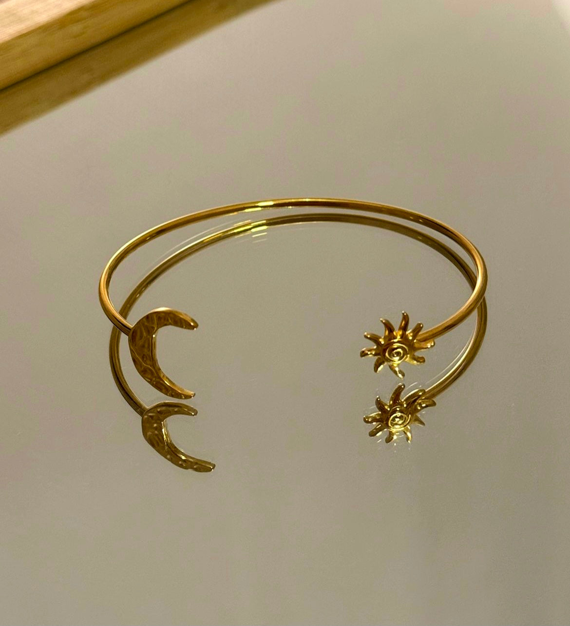 Bracelet jonc ouvert en acier inoxydable doré, présenté sur une surface réfléchissante claire. Le jonc présente un motif asymétrique à ses extrémités : un croissant de lune à finition martelée d'un côté, et un soleil rayonnant ciselé de l'autre. Le bracelet et ses motifs se reflètent nettement sur la surface en dessous.