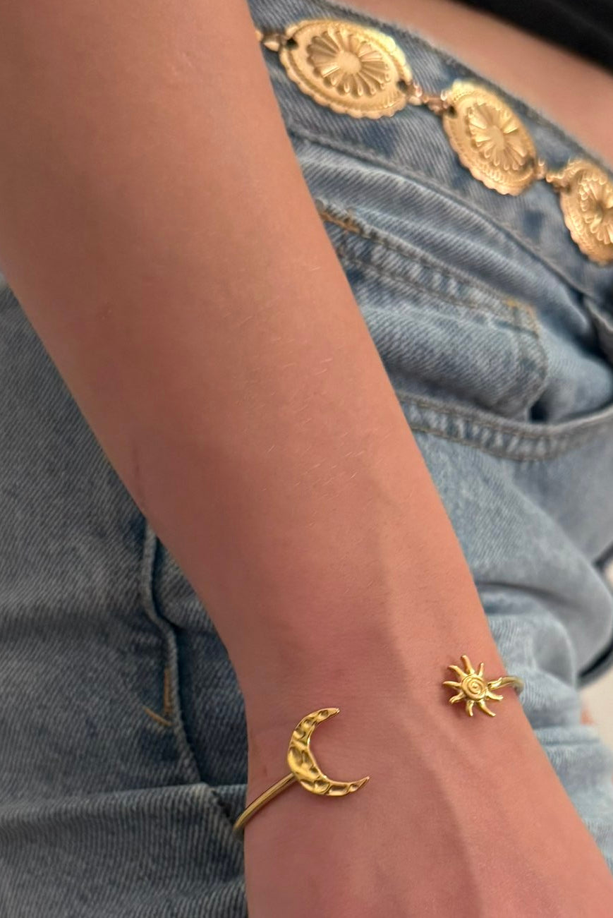 Gros plan sur le bras d'une personne portant un bracelet jonc doré ouvert, orné d'un croissant de lune et d'un soleil à ses extrémités, sur fond de jean bleu avec une ceinture dorée.